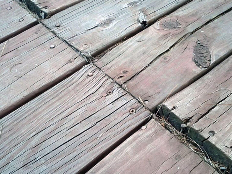 A close-up angled view of an aged wooden deck, showing cracked, cupping, and warped boards with protruding screws and a raised, uneven deck board.
