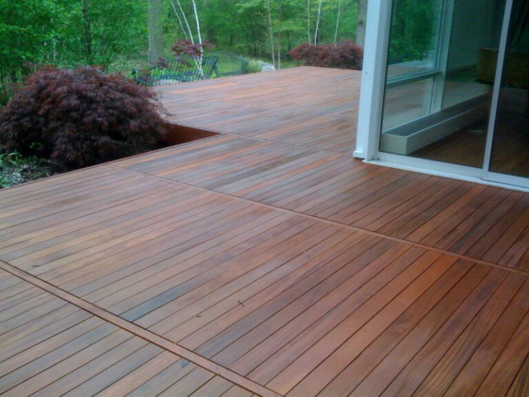 An angled view of a newly installed and caramel-stained exotic Ipe hardwood deck wrapping around a house. Beyond the deck lies a lush green backyard.
