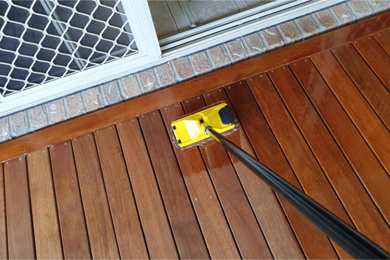 A person’s perspective looking down while staining a wooden deck with a broom-handled mop. The right side of the deck is stained a darker color, while the left side remains unstained.