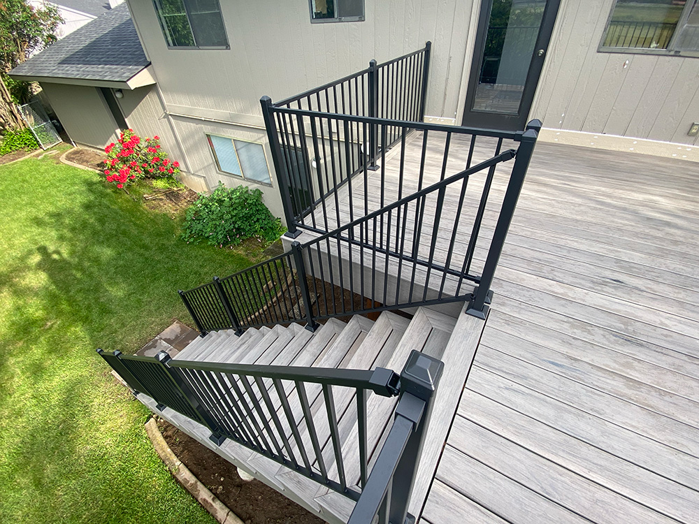 Top view of a deck staircase with handrail, decking, and fascia leading to a paver landing in a green backyard.