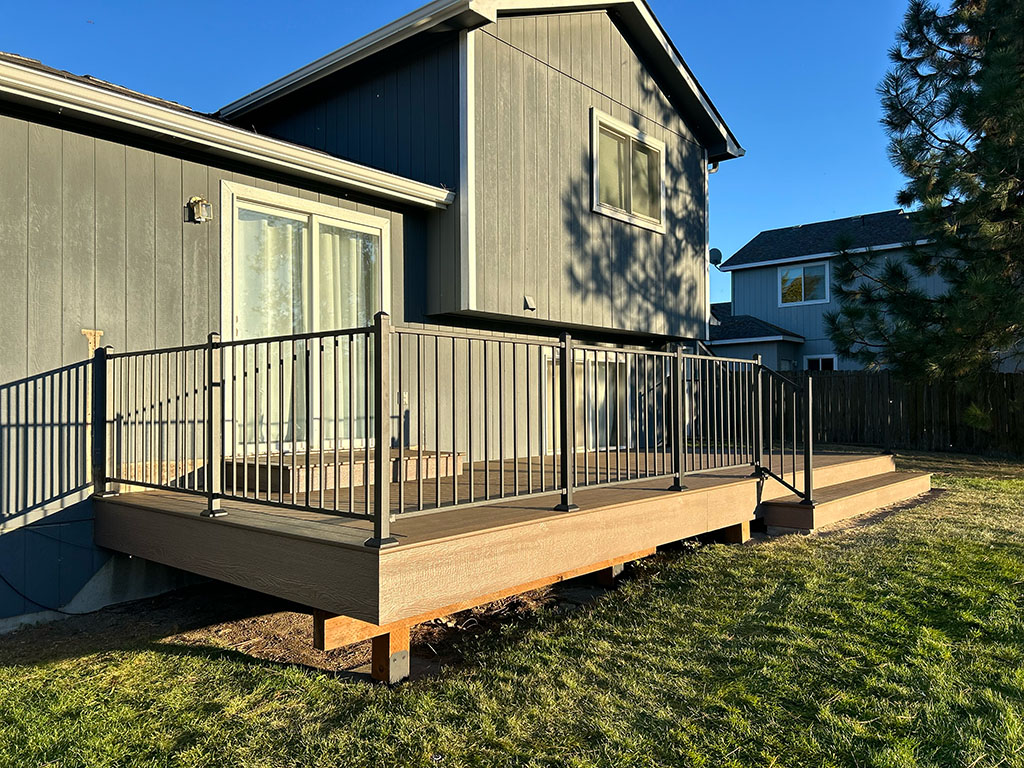 A diagonal view of a home with a freshly constructed composite deck featuring TimberTech Cocoa decking, LP wood fascia, and a Fortress Fe26 steel handrail. The left side of the deck is elevated, while the right side sits on the ground, surrounded by vibrant green grass and under a mixed blue sky with sunlight casting shadows.