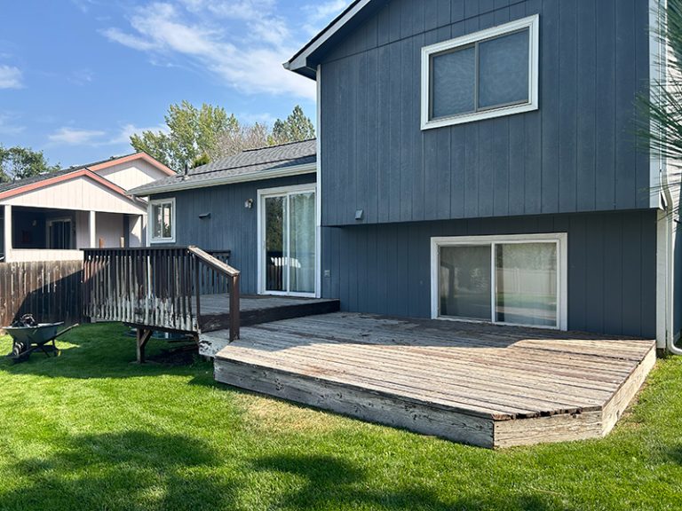 A discolored and rotting split-level wooden deck attached to a house. The right side is ground-level, while the left side with a handrail is elevated. The surrounding grass is lush and well-maintained.