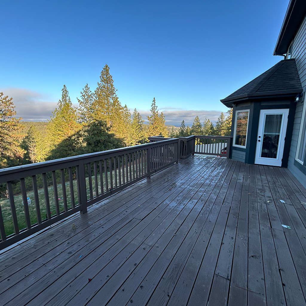 A shadowed view of an aged wooden deck with warped, rotting boards, protruding screws, and a wobbly handrail. The background features a home on the right, tall green pine trees on the left, and a dark blue sky with clouds on the horizon.
