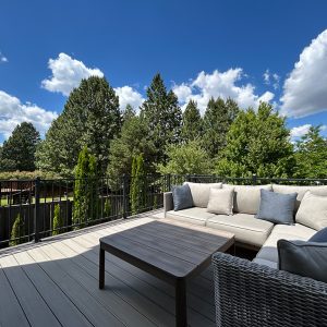 Spacious deck featuring L-shaped furniture and a coffee table, set against lush greenery and a serene sky.