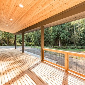 Ground-level wooden deck in the Pacific Northwest with a modern roof, tongue-and-groove soffit, wooden handrails, and metal wire accents.