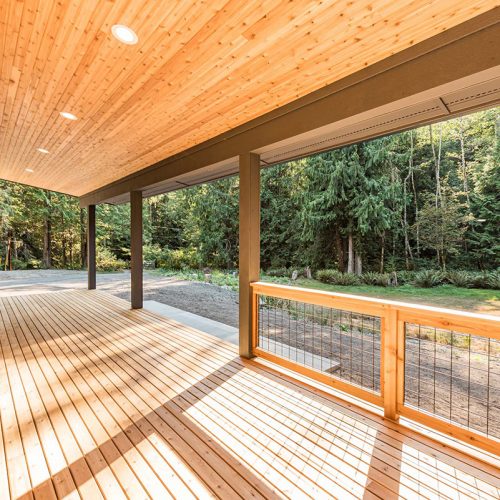 Ground-level wooden deck in the Pacific Northwest with a modern roof, tongue-and-groove soffit, wooden handrails, and metal wire accents.